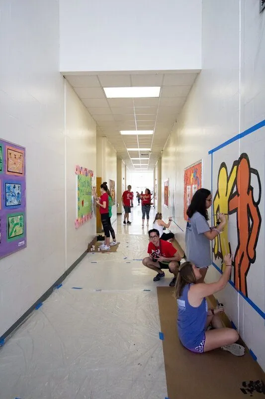 Volunteers painting bright orange dancing figures along a taped-off hallway