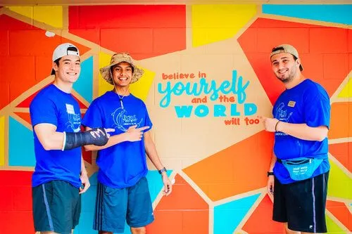 Three volunteers beside the finished mural lettered believe in yourself and the world will too