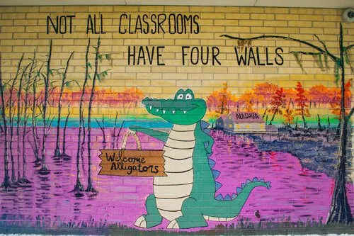 A mural of an alligator in a marsh at sunset holding a welcome sign, under hand-lettered text