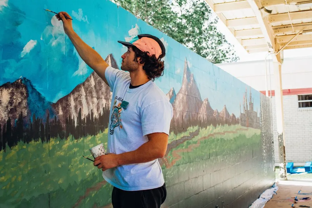A volunteer reaching up to paint a mountain landscape mural